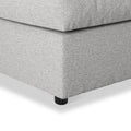A grey fabric ottoman with a visible cushion seam, resting on a black round leg; isolated on a white background.