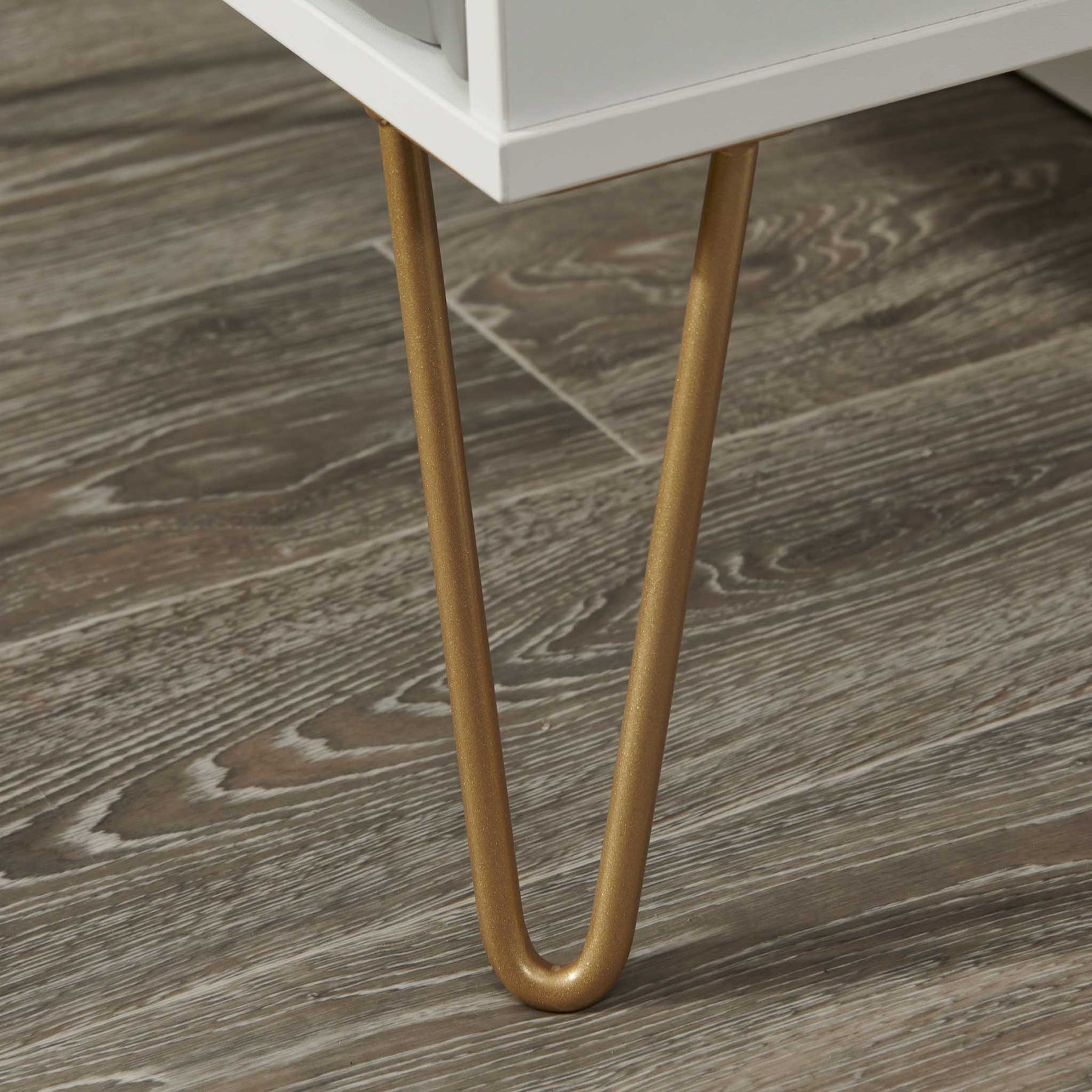 Two slender, gold-colored metal legs support a white furniture piece, against a gray wood-grain floor background.