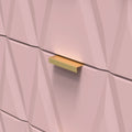 A gold-colored, rectangular drawer pull is mounted on a textured, pinkish wall with geometric patterns. There's a gentle light casting a soft shadow beneath the handle.