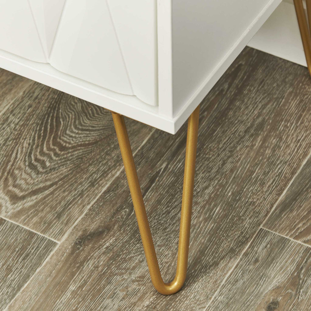 A white cabinet with a geometric design and slender, gold-colored metal legs stands on a wooden floor.